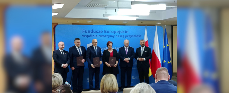 5 osób stoi na tle tablicy z symbolami Unii Europejskiej i Polskiej Flagi