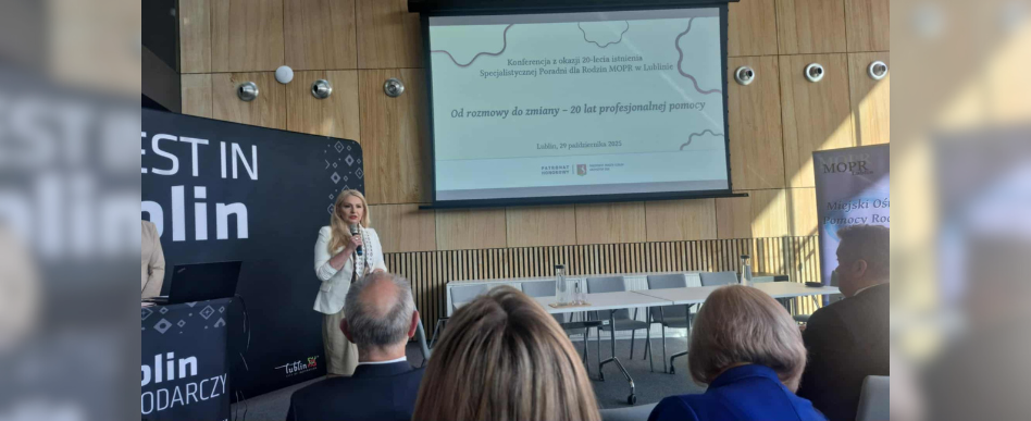 Wnętrze sali konferencyjnej. Do mikrofonu mówi kobieta. w tle biały ekran na którym wyświetlany jest napis: "od rozmowy do zmiany- 20 lat profesjonalnej pomocy"