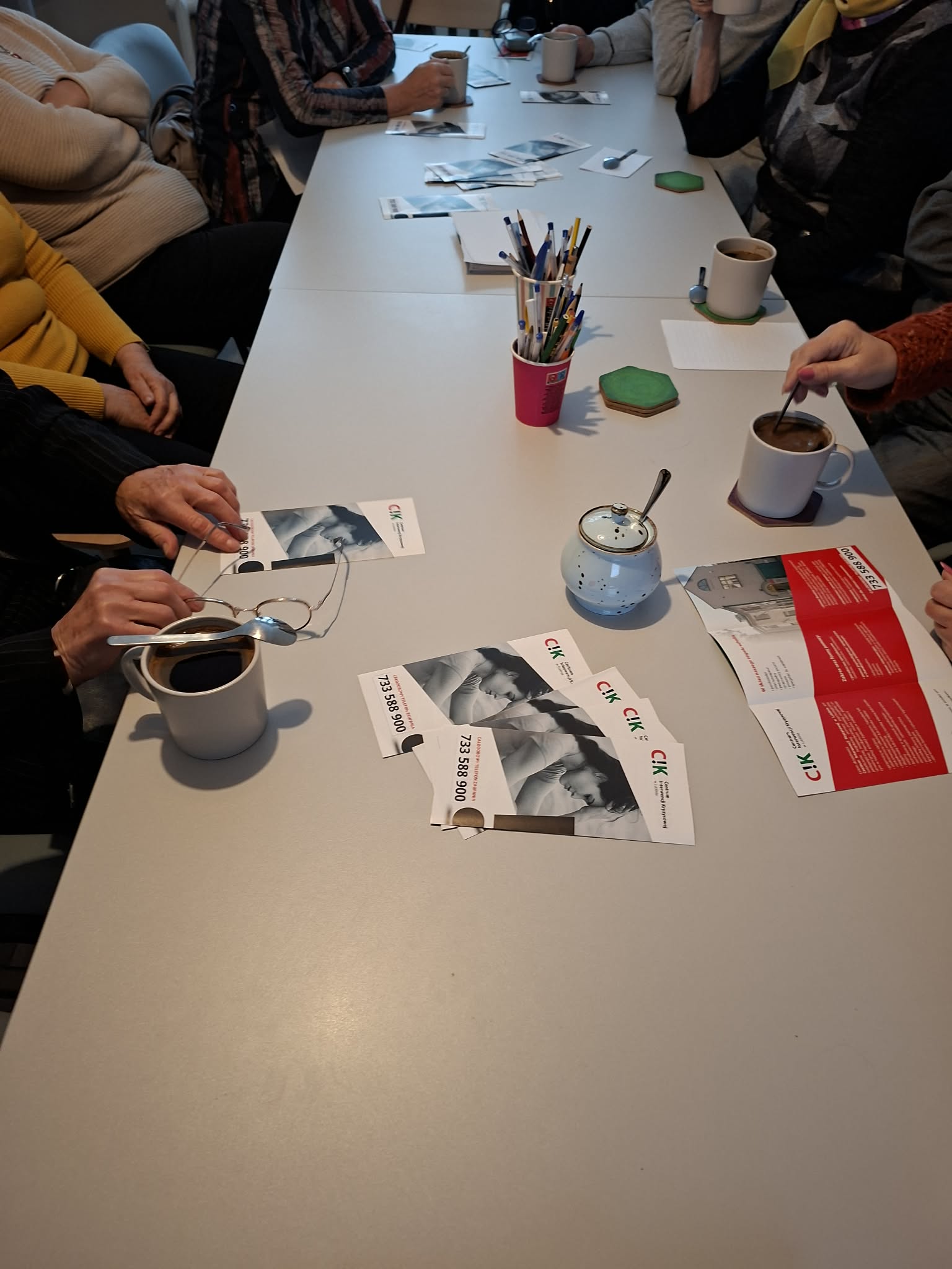 widok z góry na blat stołu, na którym leżą ulotki Centrum Interwencji Kryzysowej, kubki z napojami, widoczne są ręce osób siedzących przy stole