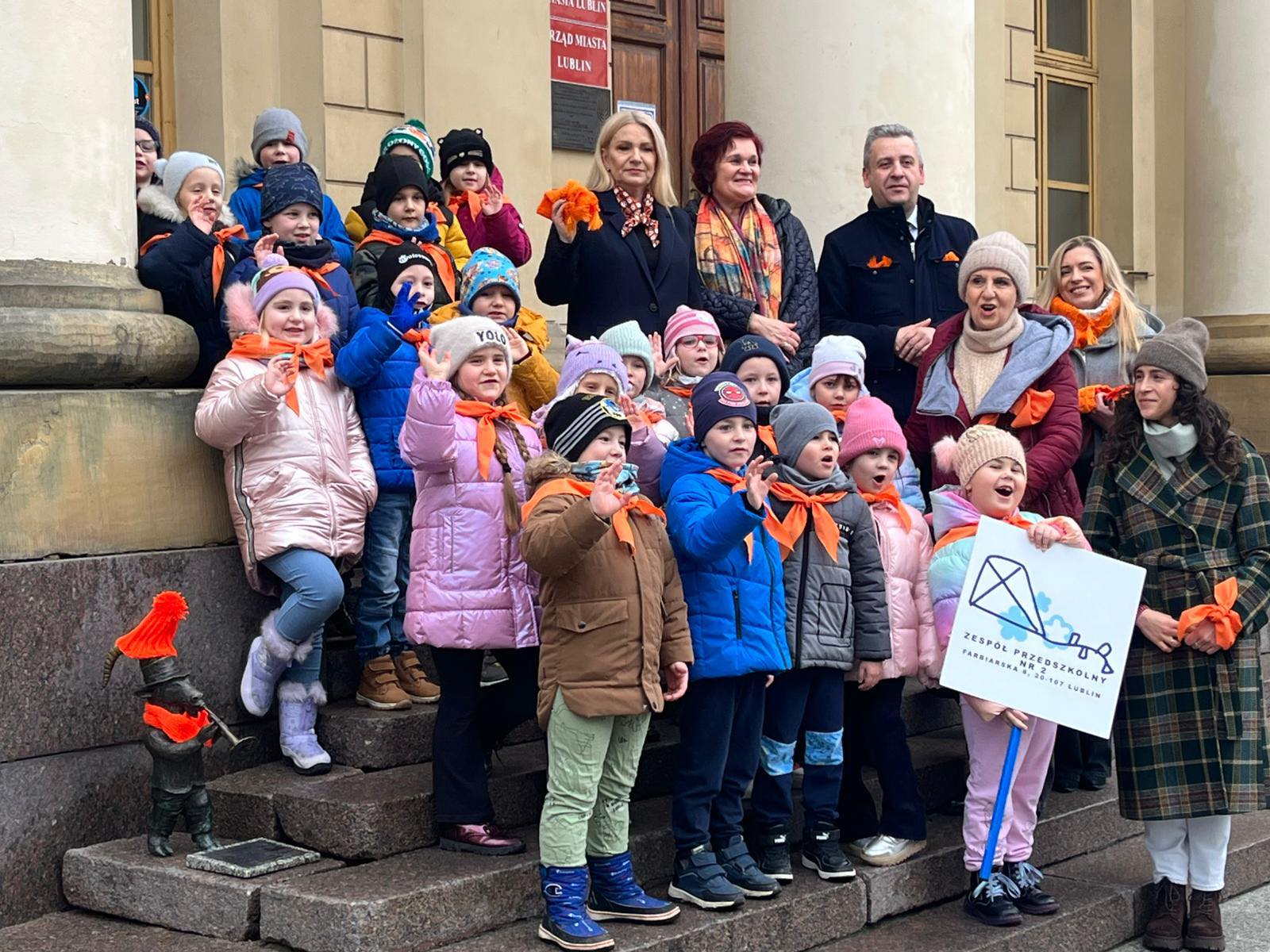 grupa dzieci i dorosłych ubranych w pomarańczowe szaliki stoją na schodach budyn,u