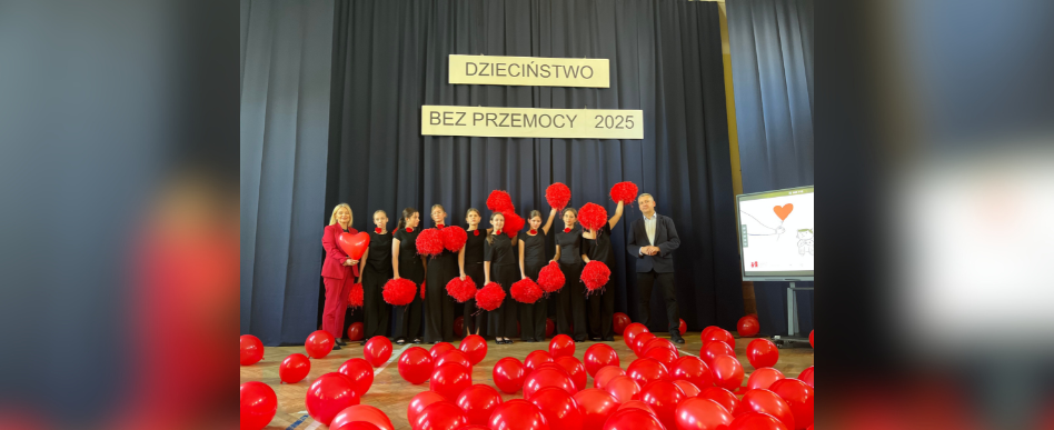 wnętrze sali gimnastycznej. Akademia, wiele osób pozuje do zdjęcia z czerwonymi pomponami i balonami. Z tyłu, na zasłonie napis "dzieciństwo bez przemocy 2025"