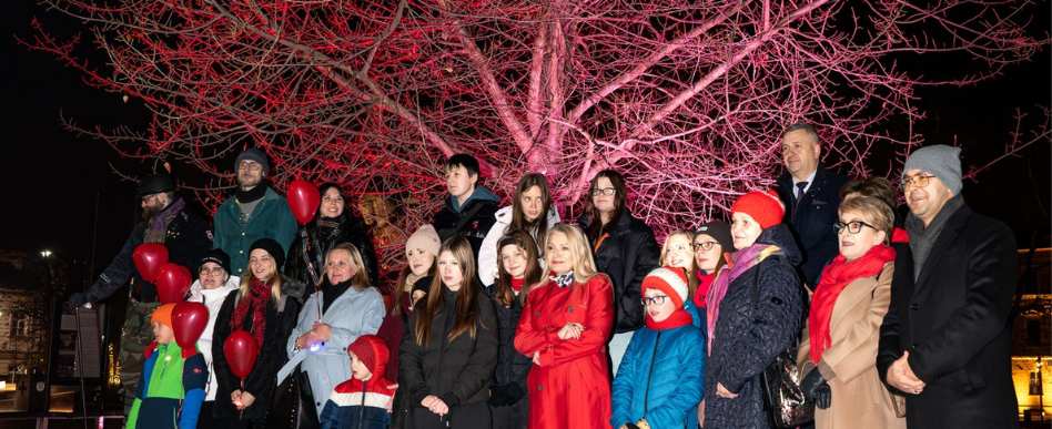 grupa osób pozujących do zdjęcia na placu litewskim w Lublinie na tle drzewa oświetlonego na czerwono. Osoby trzymają czerwone balony i mają elementy czerwonych ubrań